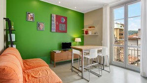 A room with a green wall, a wooden desk with a chair, a white sofa, a television on a wooden cabinet, and a balcony with a view of buildings. - Delightful Holiday Apartment Steps from the Port with Mountain Views (como)