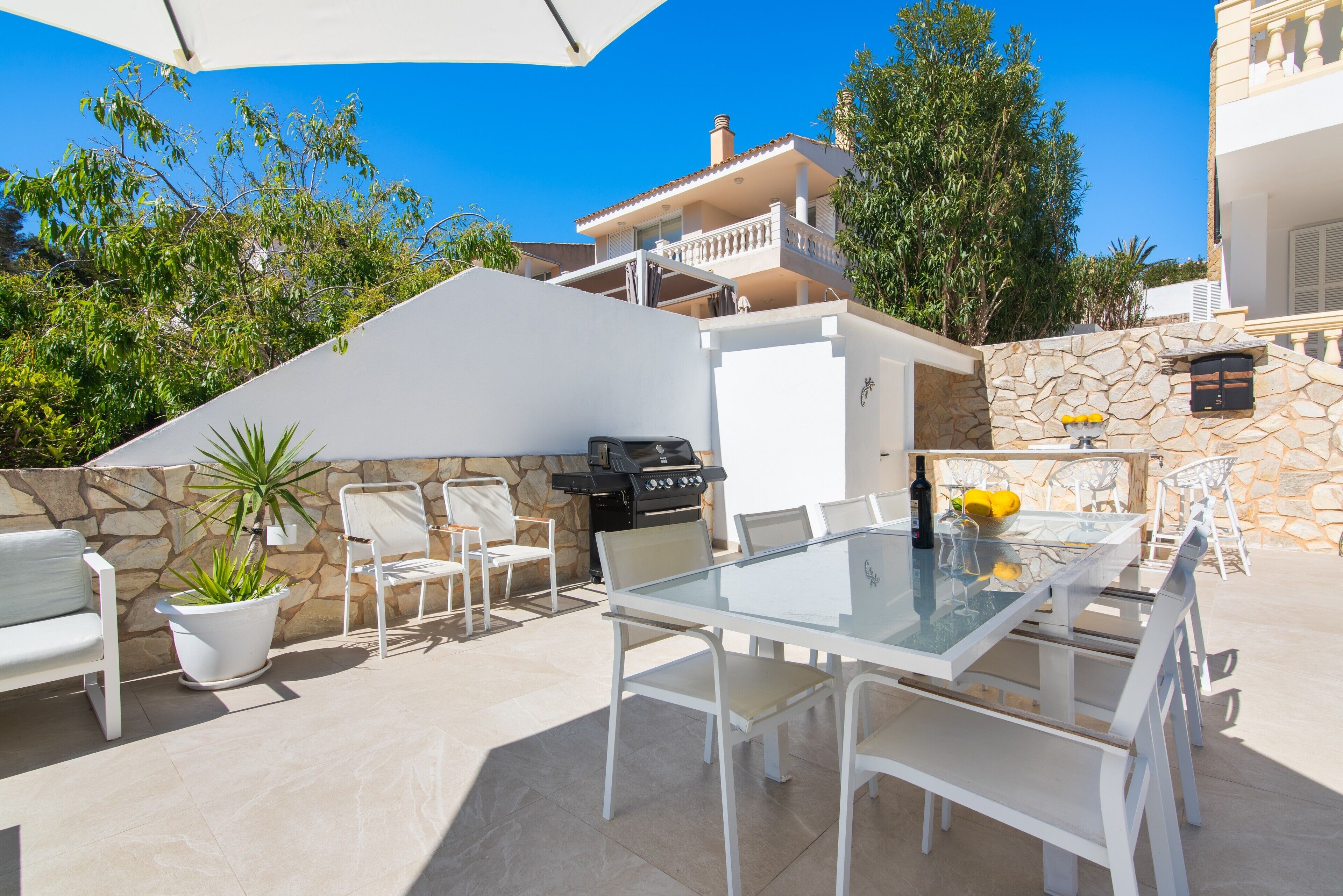 Une terrasse extérieure avec une table en verre, des chaises blanches, un barbecue et une plante en pot.