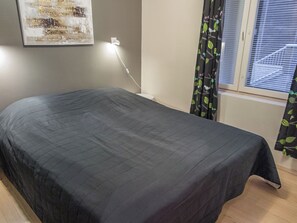 A neatly made bed in a room with a window, a wall-mounted light, and a piece of abstract art.