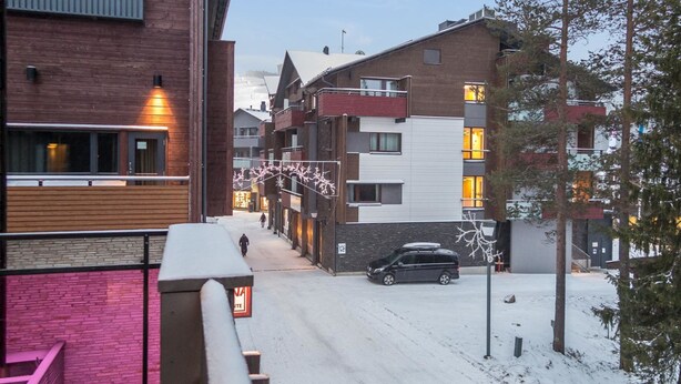 A snow-covered street with multi-story buildings, a parked car, and a person walking.