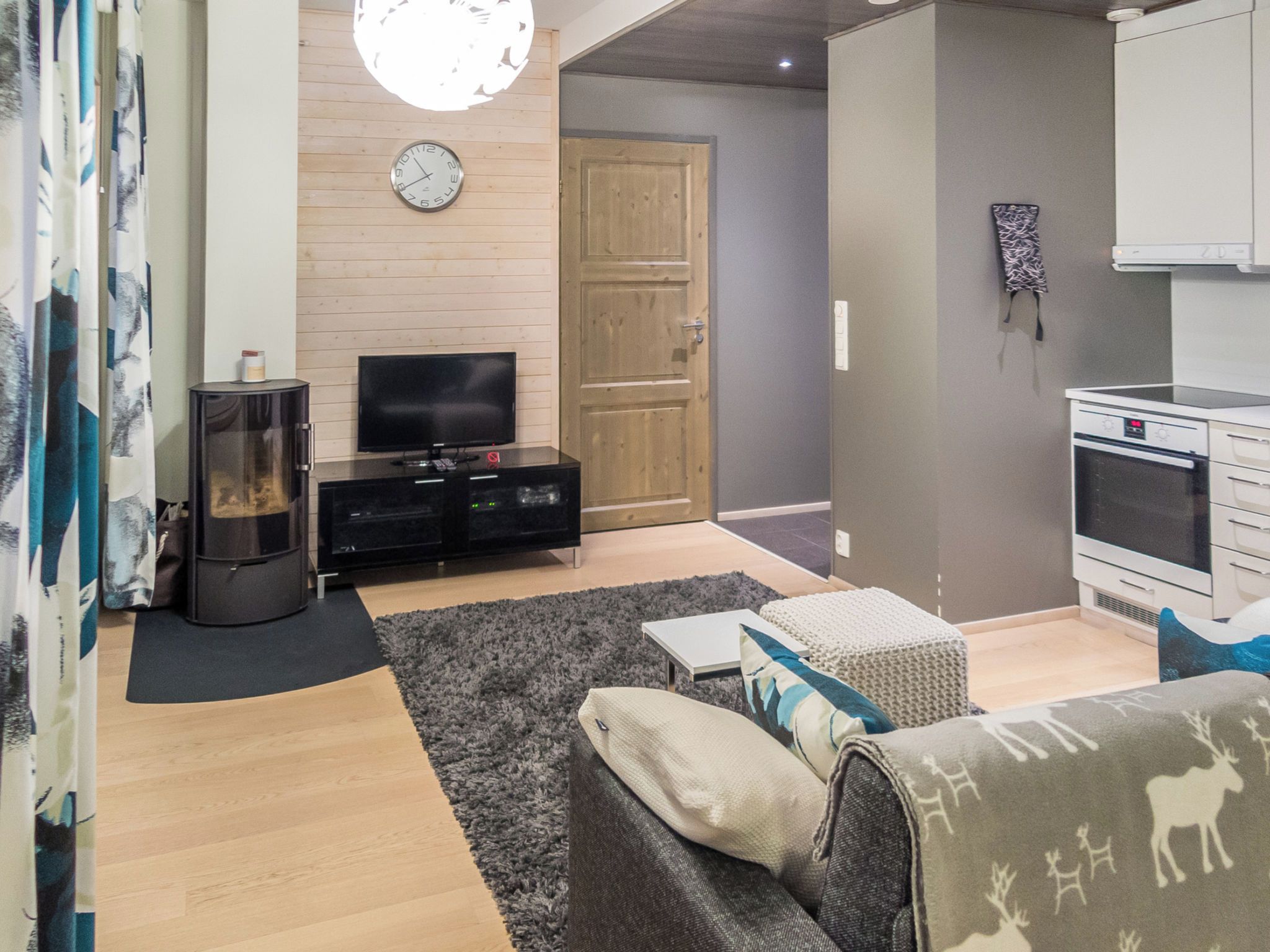 A living room with a television, sofa, and a fireplace. There is a wooden door and a kitchen area with a stove.