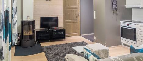A living room with a television, sofa, and a fireplace. There is a wooden door and a kitchen area with a stove.