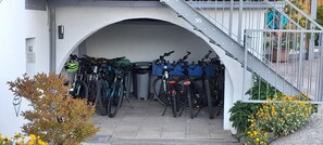 Katettu pyöräparkkialue, jossa on useita pyöriä, roskakori ja portaikko, joka johtaa ylemmälle tasolle.
