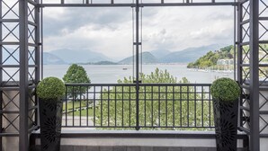 Ein Balkon mit Metallgeländer, zwei Blumentöpfen und Blick auf ein Gewässer mit Bergen im Hintergrund.