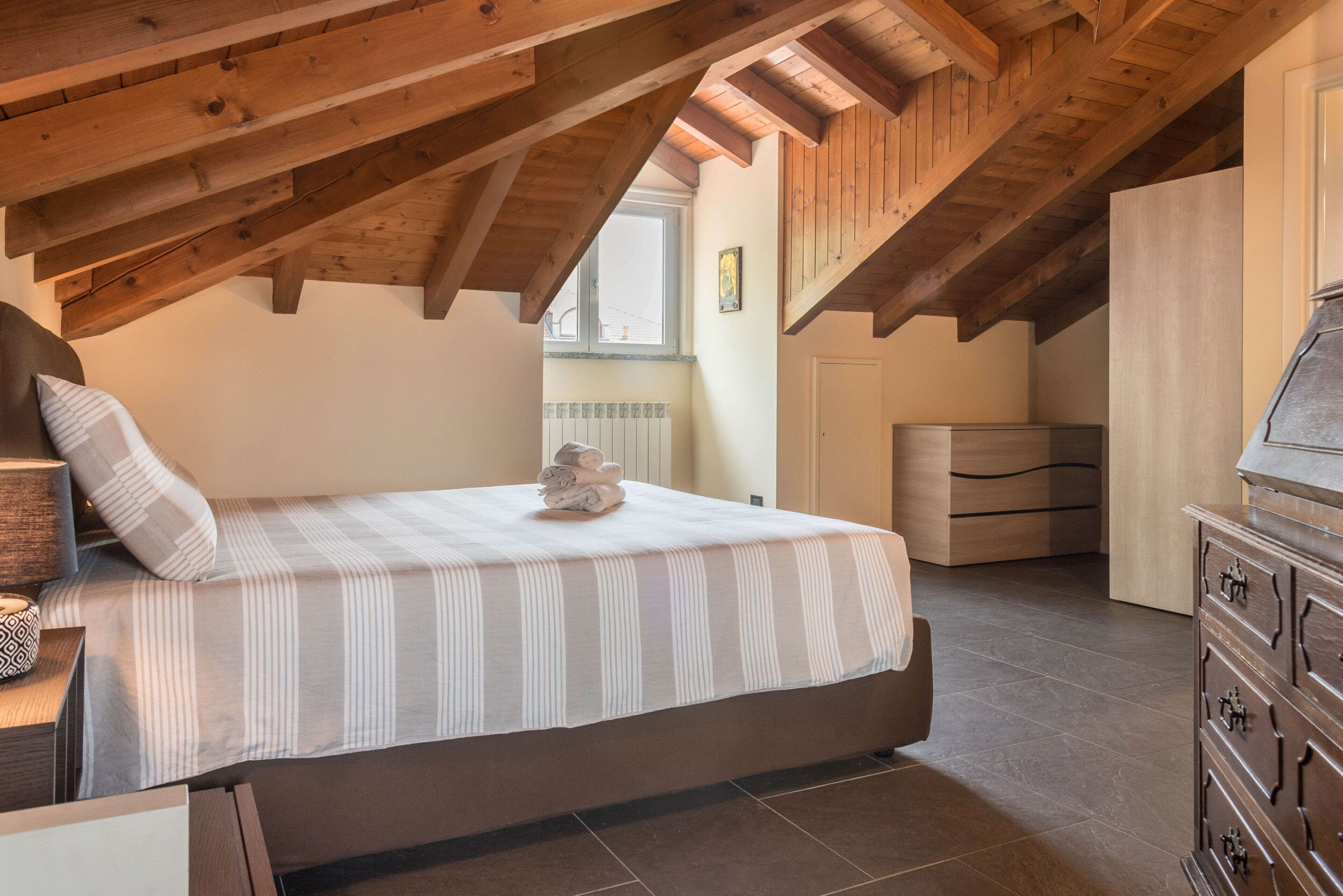 A bedroom with a bed, wooden ceiling beams, a dresser, and a window.