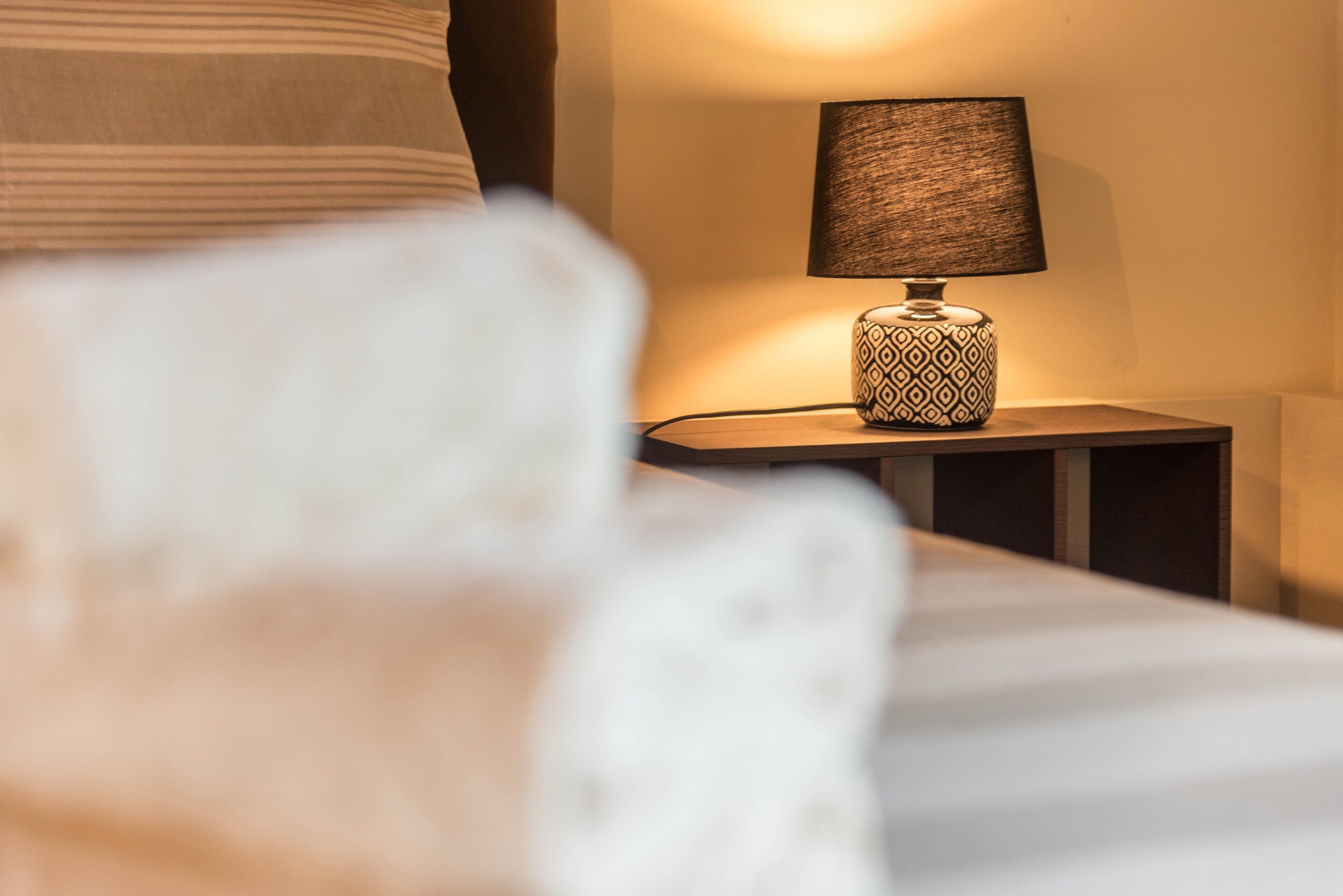 A neatly made bed with a pillow, a bedside lamp with a patterned base, and a wall-mounted lamp.