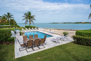 A pool area with lounge chairs and a dining table, surrounded by greenery and a view of the sea.