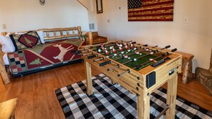 A cozy room with a foosball table, a sofa with a deer-themed blanket, and a wall-mounted American flag. - Ridgetop Lakeview - Bedroom Cabin on Lake Nantahala w/ Private Deck (Topton)