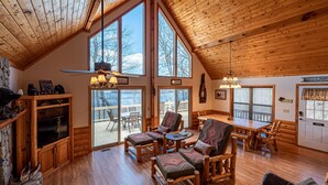 Ruim interieur van een chalet met hoge plafonds, grote ramen, een houten eethoek en een stenen open haard.