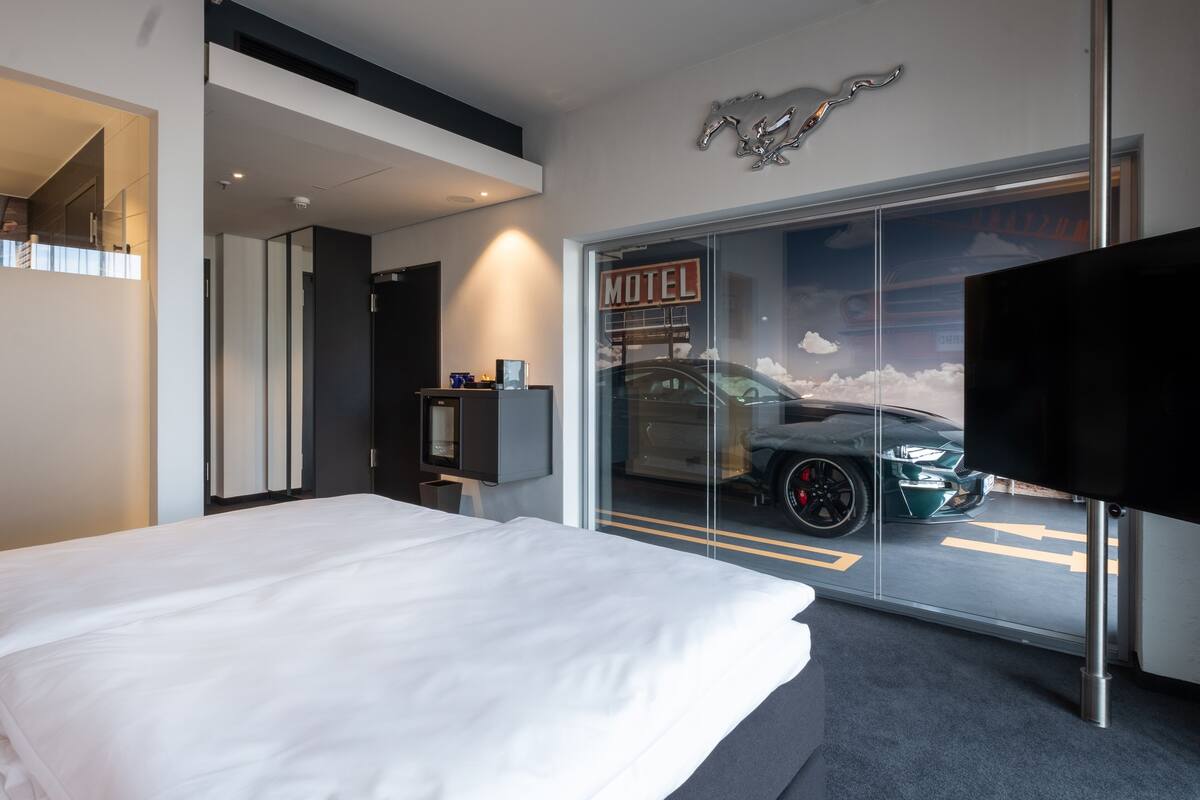 Quarto de hotel moderno com uma cama grande, uma televisão, uma porta de vidro que dá para um estacionamento com um carro e um letreiro com a inscrição 'Motel'.