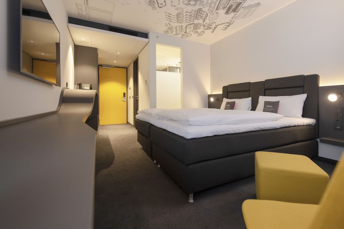 Quarto de hotel moderno com uma cama grande, mesa de cabeceira, uma cadeira e um design de teto singular.