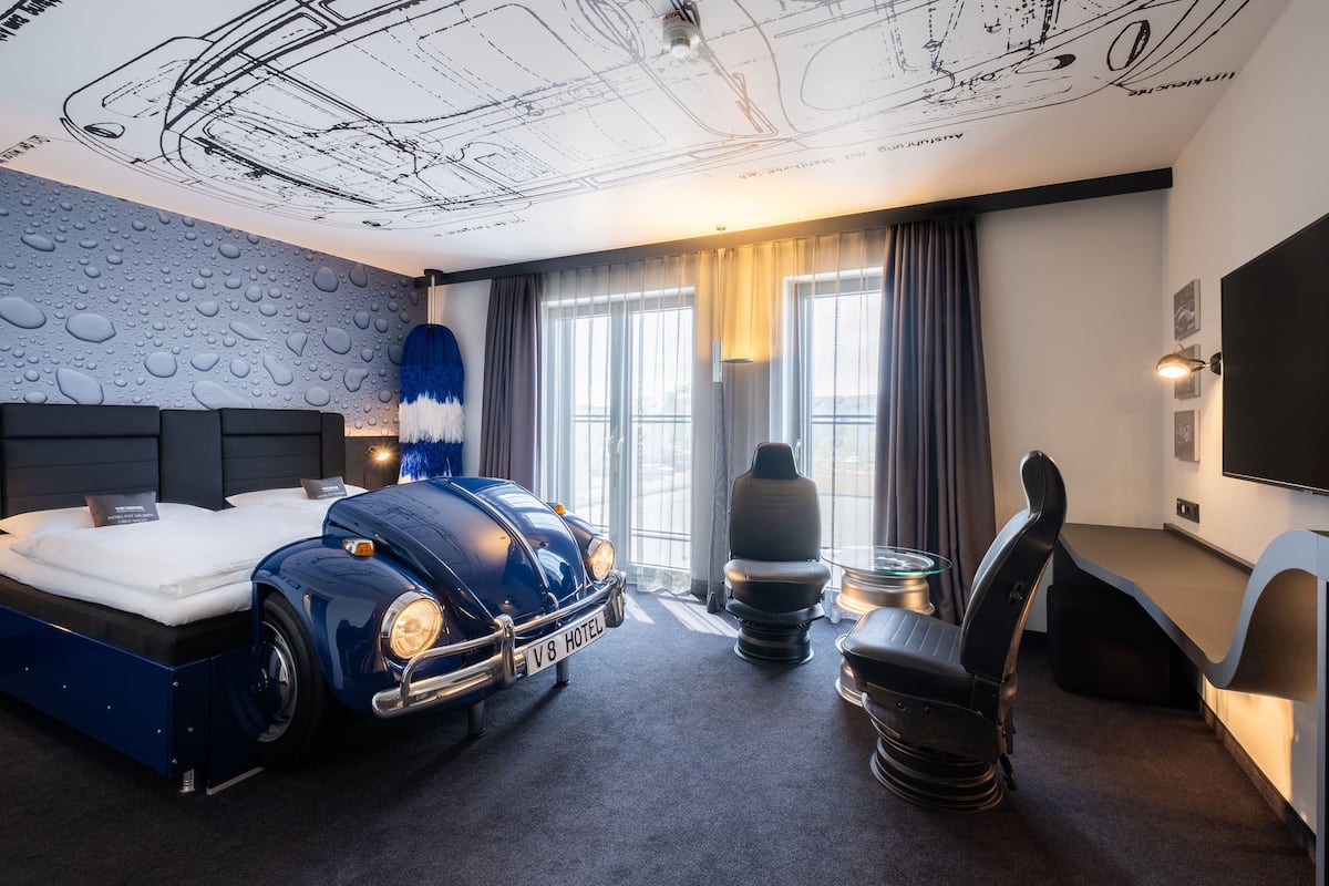 Quarto de hotel moderno com uma cama, uma cama em formato de carro azul exclusivo, uma escrivaninha e uma televisão grande.
