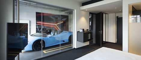 A modern hotel room with a large bed, a glass wall with a car reflection, a desk, and a TV.