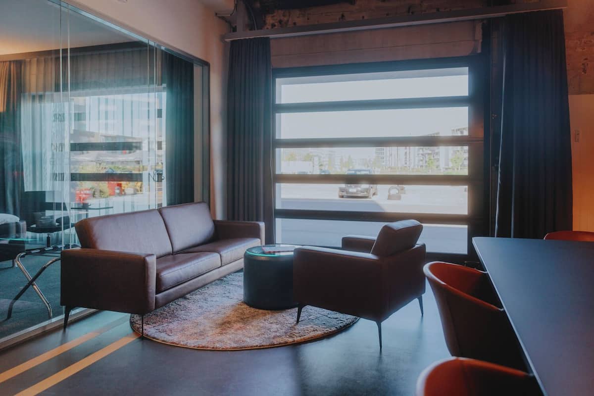 Uma sala de estar moderna com um sofá de couro, uma mesa de centro redonda e uma mesa de jantar. Grandes janelas oferecem vista para a rua.