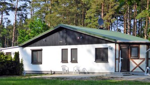 A single-story building with a green roof, white exterior walls, and a black door. There are three windows and a satellite dish on the roof. - Ferienwohnung 1 - Ferienhaus am Waldessaum (Fuhlendorf)