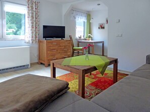 A living room with a sofa, a wooden table with a green tablecloth, a TV on a wooden cabinet, and a chair.