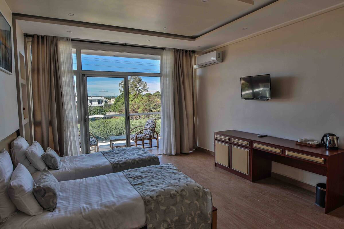 A hotel room with two beds, a desk, a TV, and a view of the outdoors.