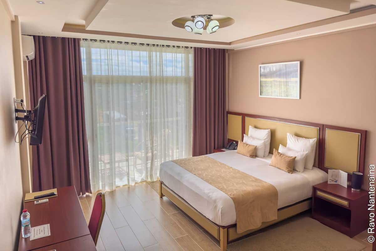 A hotel room with a large bed, a bedside table, a desk with a chair, a TV mounted on the wall, and a window with curtains.