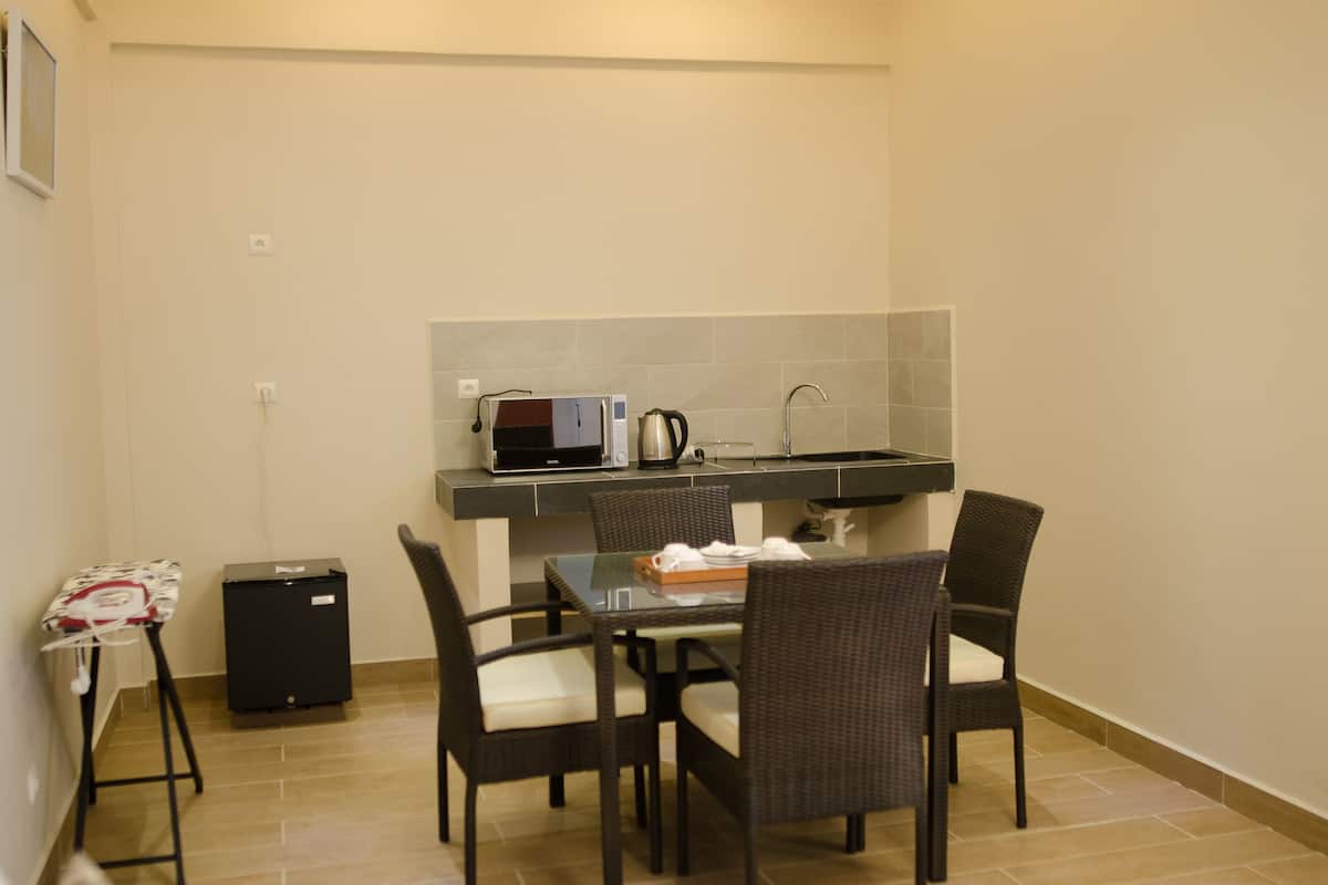 A compact kitchen with a microwave, kettle, and sink. There are four wicker chairs around a small table. A compact refrigerator and ironing board are also visible.