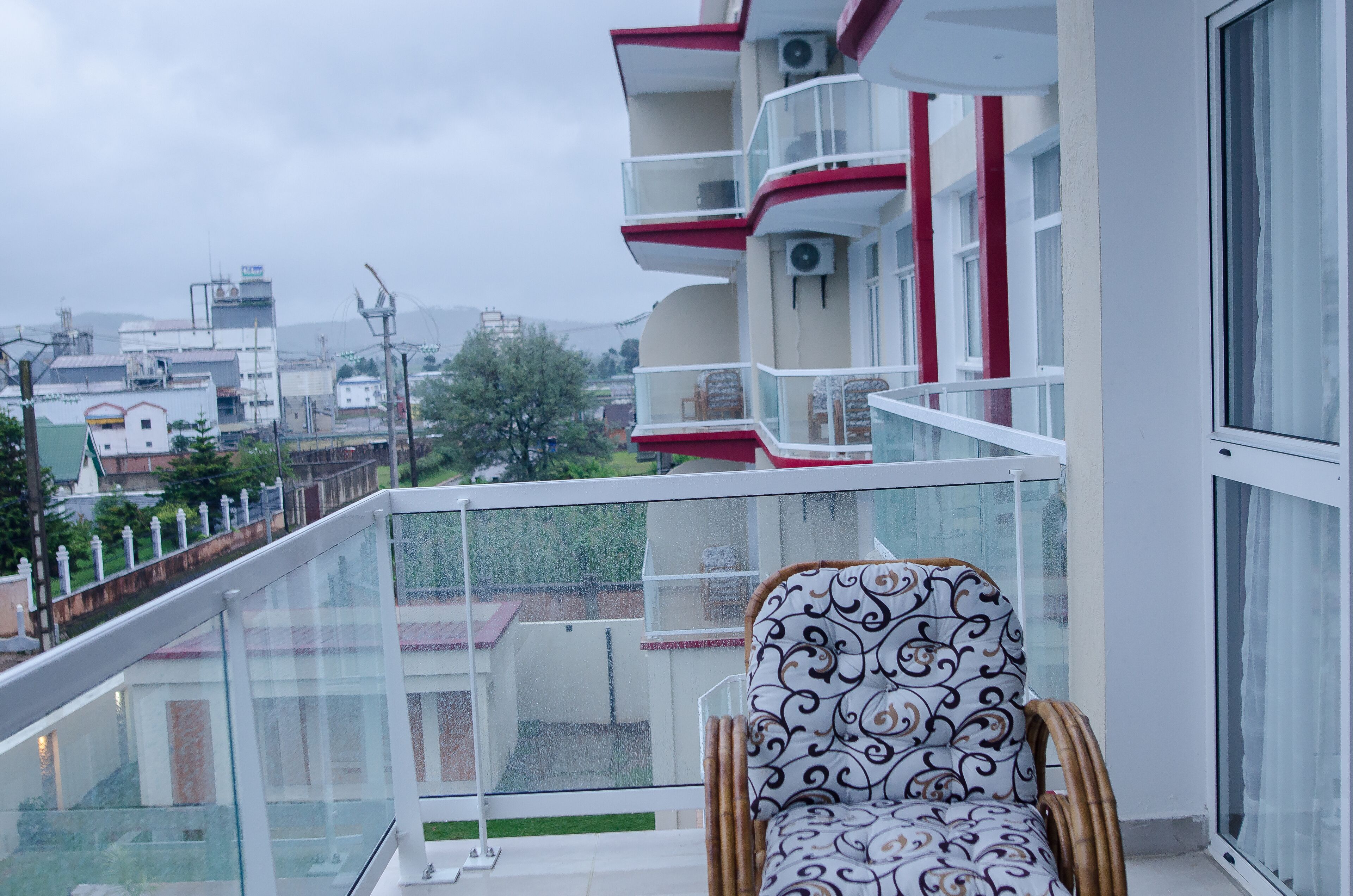 Un balcón con una silla de mimbre, barandilla de vidrio y vistas a edificios y árboles.