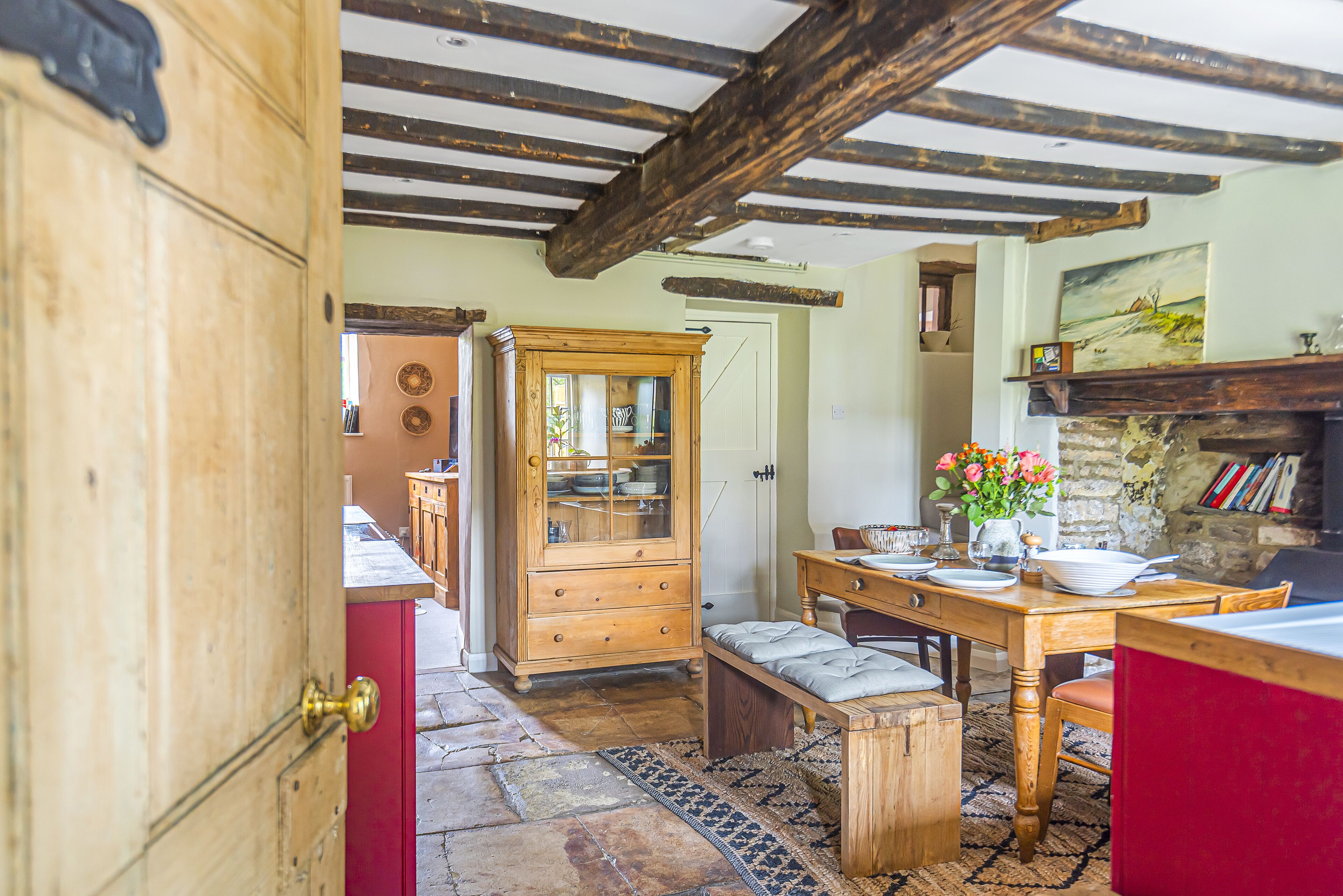 A rustic kitchen with exposed wooden beams, a wooden cabinet, a dining table with chairs, and a red cabinet.