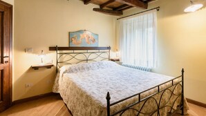 A bedroom with a bed, wooden ceiling beams, a nightstand with a lamp, a painting on the wall, and a window with curtains. - Borgo dei Corsi - RIBUSTINO 2 Apartment (Raggiolo)