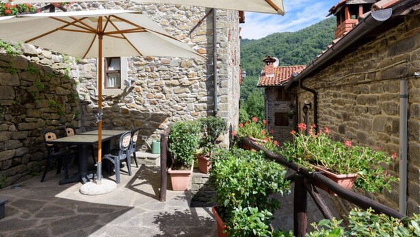 A stone building with a patio area featuring a table and chairs under a large umbrella, surrounded by potted plants and a stone wall. - Borgo dei Corsi - RIBUSTINO 2 Apartment (Raggiolo)