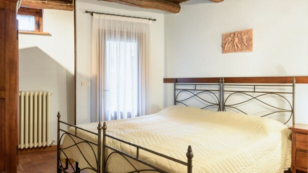 Un dormitorio con estructura de cama metálica, cabecera de madera, radiador, ventana con cortinas y una escultura en la pared.