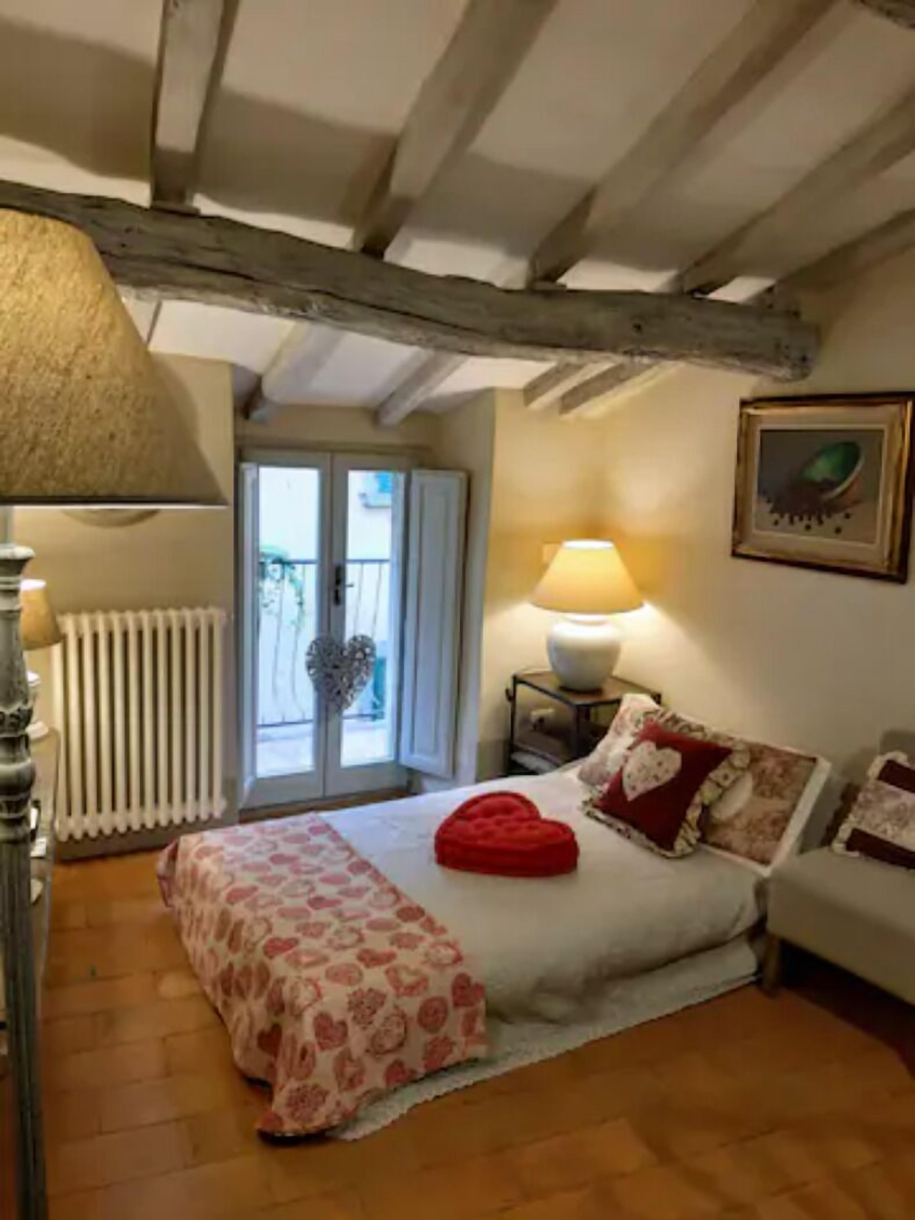 A cozy bedroom with a wooden ceiling, a bed with a red heart-shaped pillow, a bedside table with a lamp, and a window with a heart decoration.