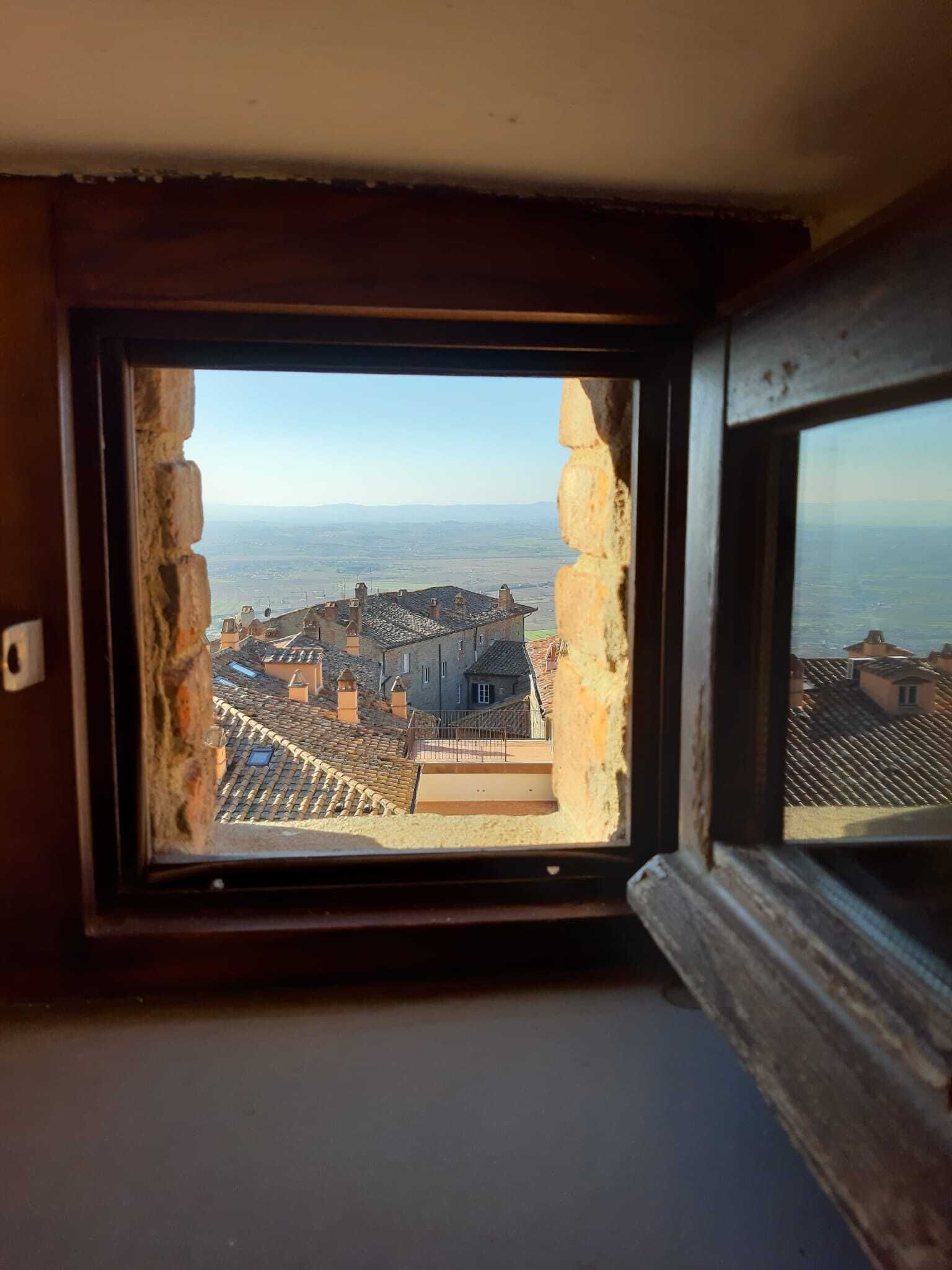 A window with a view of a coastal town, featuring terraced rooftops and a distant sea.
