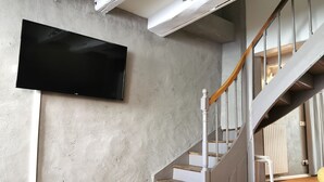 Moderni olohuone, jossa on seinään kiinnitetty litteänäyttöinen televisio, betoniin tehty seinä, puinen portaikko ja valkoinen sohvapöytä, jonka päällä on kaukosäädin.