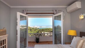 Une chambre à coucher avec un lit, une table de chevet, une lampe et une vue sur le paysage urbain, visible à travers une porte ouverte.