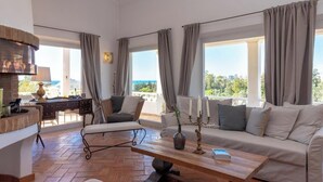 Un vaste espace de vie comprenant un canapé, un fauteuil et une table basse, un bureau avec une lampe, ainsi que de grandes fenêtres équipées de rideaux.