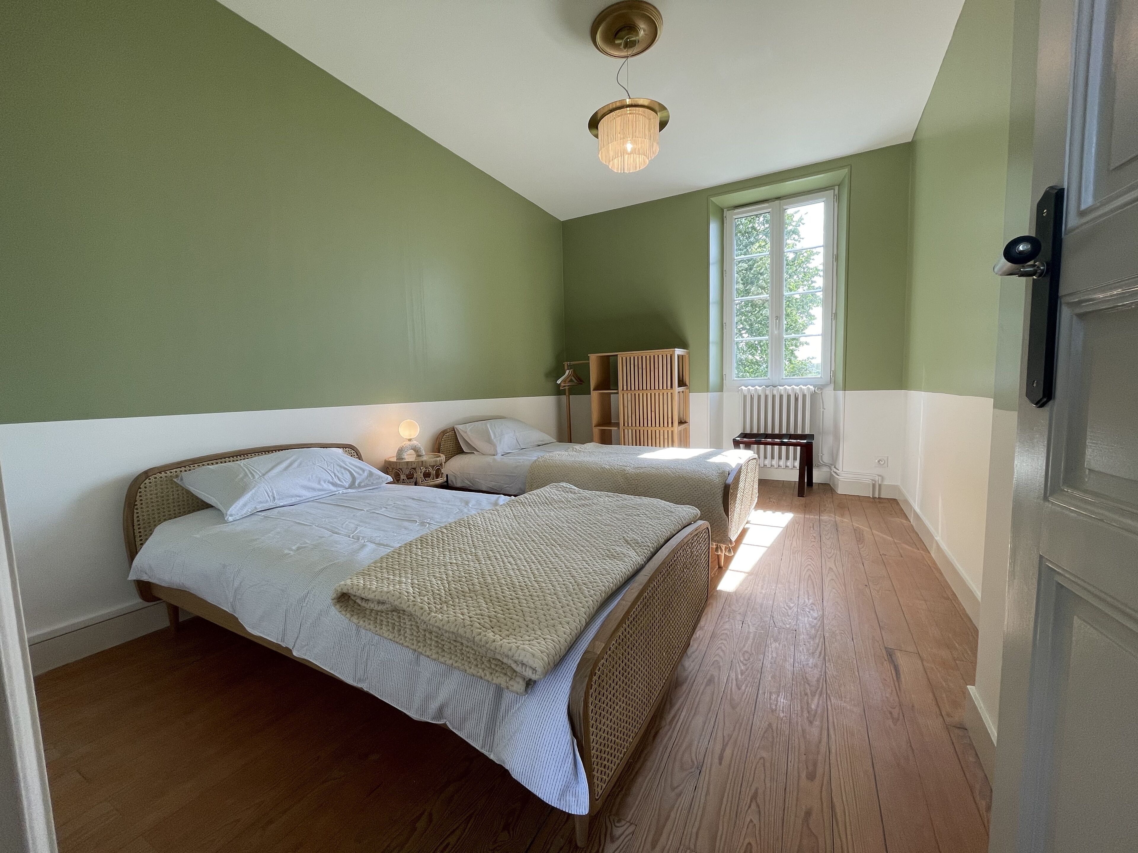 A bedroom with two beds, a wooden floor, a radiator, and a window with a view of trees.