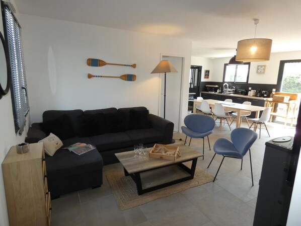 Un salon moderne avec un canapé noir, une table basse en bois et des chaises bleues. On aperçoit une cuisine à l’arrière-plan, avec une table à manger et des chaises.