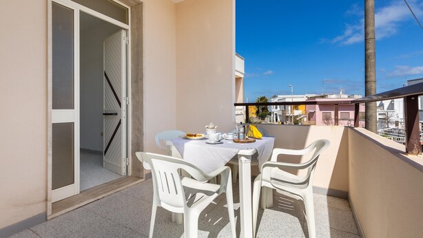 A balcony with a table set for two, chairs, and a view of buildings in the background. - Carlotta`s House by BarbarHouse (San Foca)