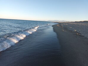 Una playa con olas que acarician suavemente la orilla, cielo despejado y montañas a lo lejos.