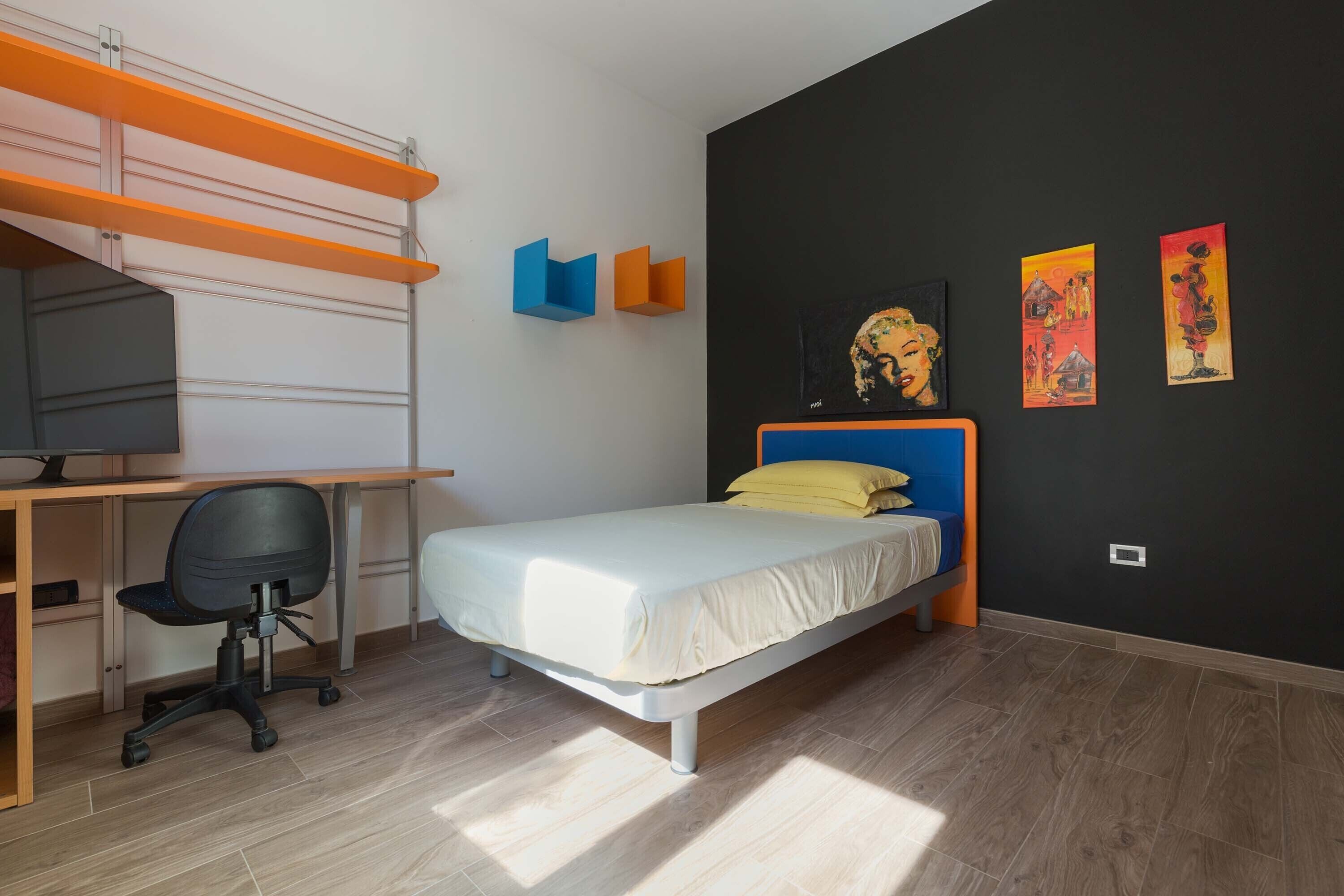 A bedroom with a bed, a desk, and a chair. There are wall art pieces and a shelf with orange and blue objects.