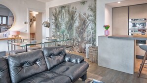 Ein modernes Wohnzimmer mit einer Ledersofa, einem Glastisch, einer Küchenzeile mit Bar und einem tropischen Wandgemälde.