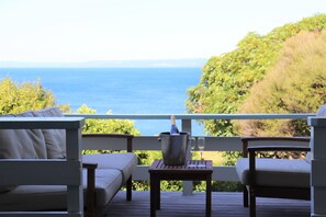 Eine Holzterrasse mit einem weißen, gepolsterten Sofa und Sesseln, einem kleinen Tisch mit einer Flasche und einem Glas sowie Blick aufs Meer.