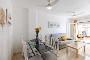 A modern living room with a glass dining table, white chairs, a grey sofa with colorful cushions, a small wooden coffee table, and a ceiling fan. - Fidalsa Bay View Romance (Guardamar)
