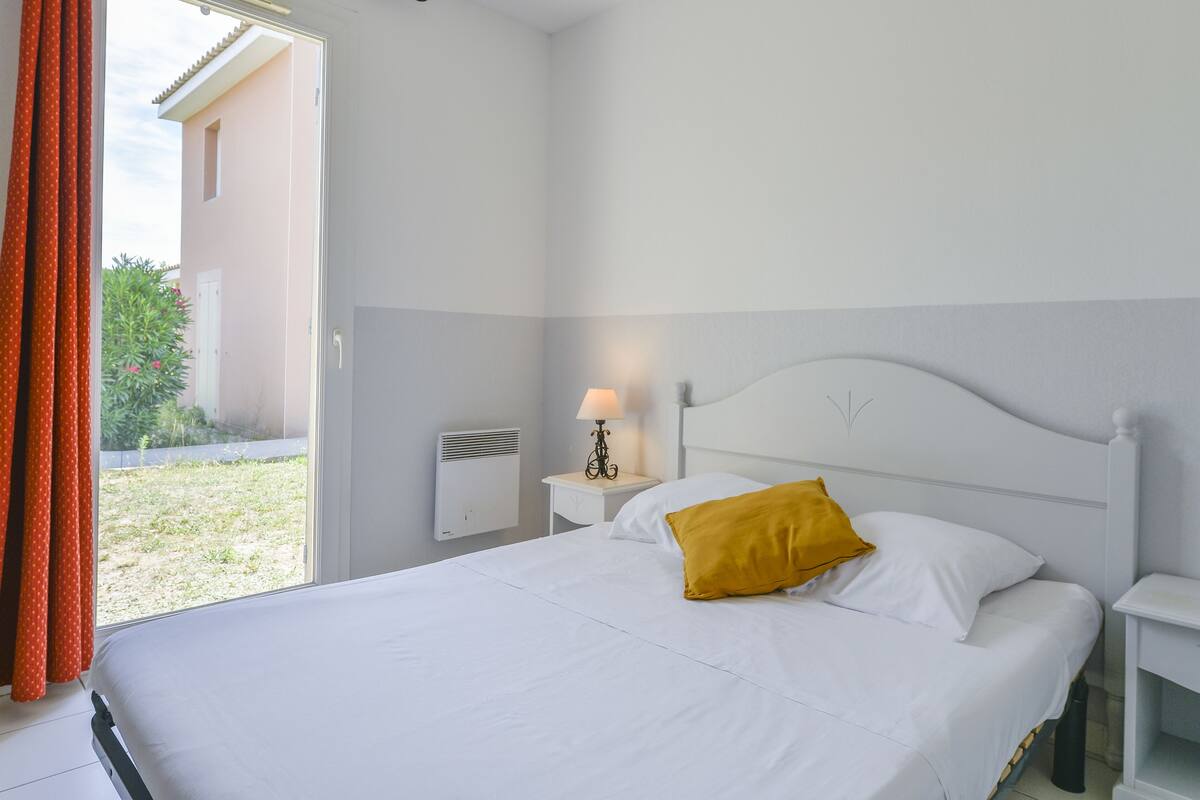 Een slaapkamer met een bed, nachtkastje, lamp en raam met gordijnen.