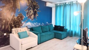 A living room with a sectional sofa, a white armchair, a teal accent wall, and a ceiling-mounted air conditioner. - Luxury 2-Bedroom Apartment in MiniSmart Complex RainbowGarden (Balchik)