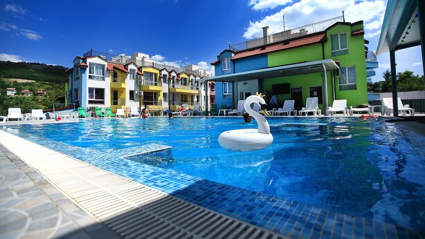 A swimming pool with a swan float, surrounded by colorful buildings and lounge chairs. - Luxury 2-Bedroom Apartment in MiniSmart Complex RainbowGarden (Balchik)
