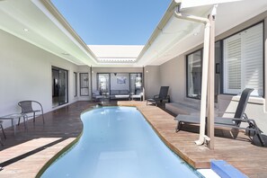 Un área de piscina interior moderna con una terraza de madera, sillones de descanso y un muro de cristal que da a una sala de estar.