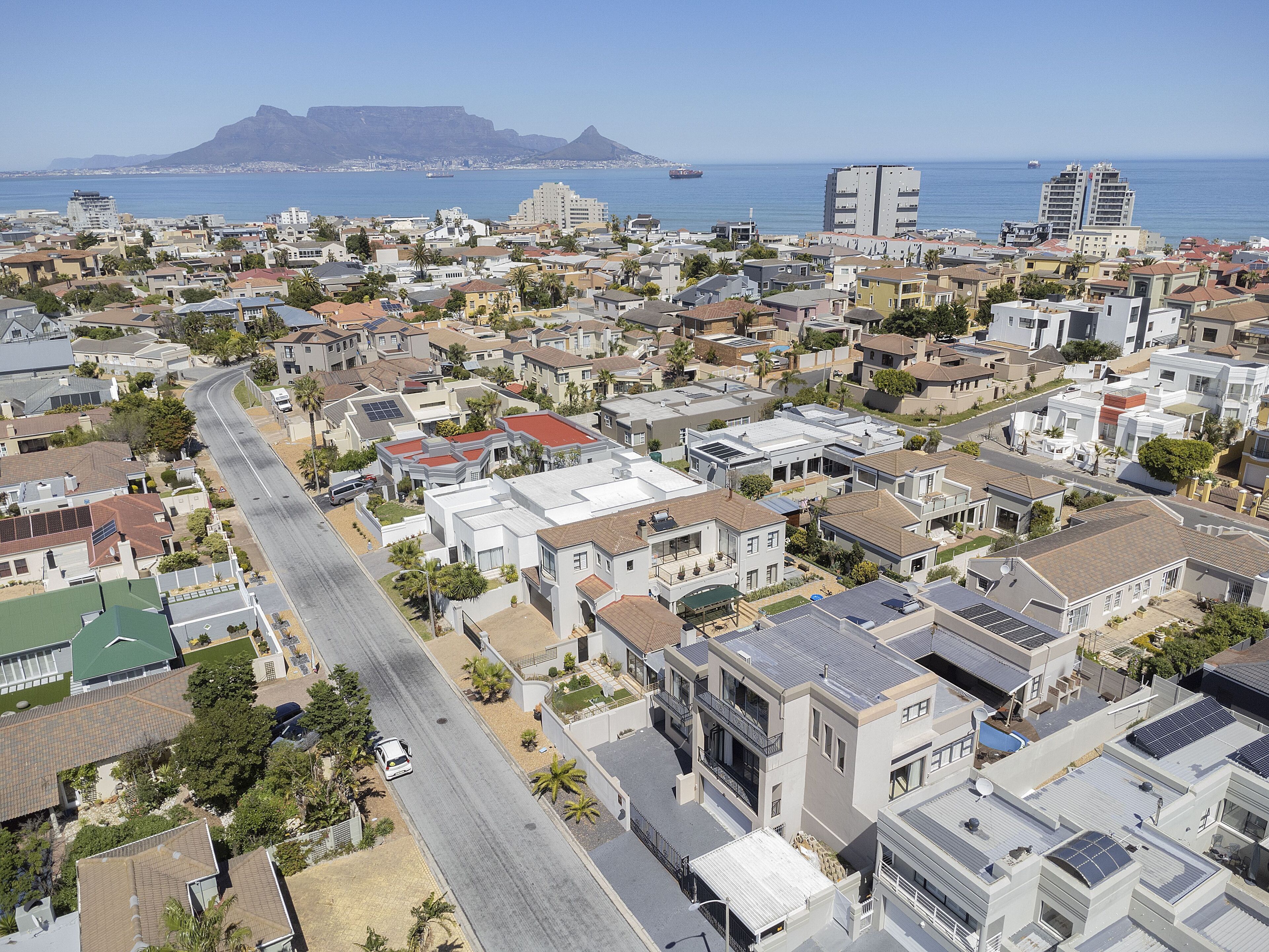 A residential area with a main road, houses with solar panels, and a view of the ocean and mountains in the distance.