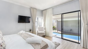 A bedroom with a large bed, a flat-screen TV, a chair, and a sliding glass door leading to a pool area. - Villa Balmoral 14 (Cape Town)