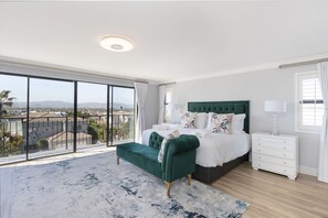 A spacious bedroom with a large window, a green upholstered bench, a white bed with decorative pillows, and a white nightstand. - Villa Balmoral 14 (Cape Town)
