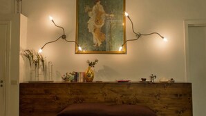 Une chambre avec une tête de lit en bois, un tableau au mur, un vase rempli de fleurs et une étagère avec des livres et une cafetière.