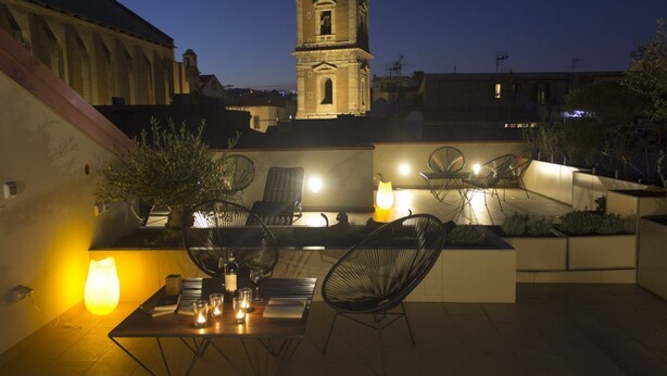 Un patio al aire libre con una mesa dispuesta para comer, rodeada de sillas y una lámpara. Se ve una torre del reloj al fondo.